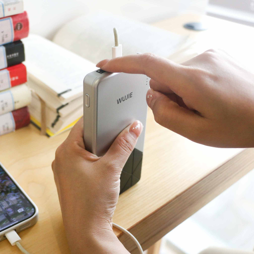 A hand charges a phone using a “WUJIE” power bank, with books resting nearby. The scene reflects how technology quietly supports our daily study and mobility, ensuring our connection to the world remains powered and present.